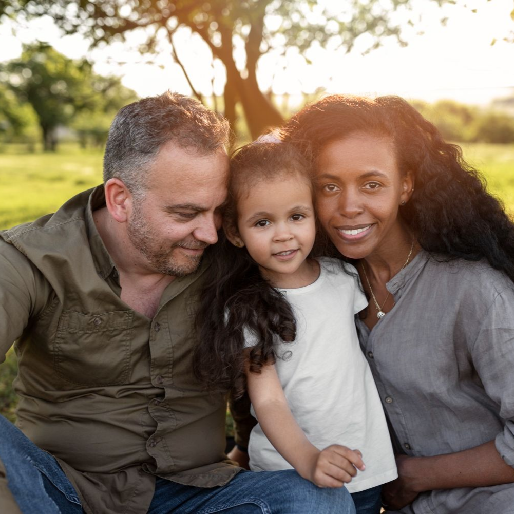 Family sitting on the floor smiling at a toddler. Mom holding child. Dad holding child's hand. Living room setting.