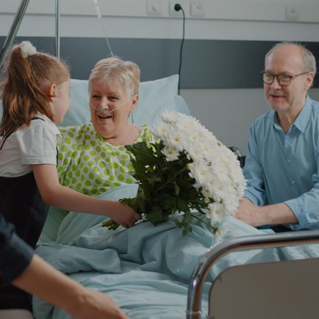 Family sitting on the floor smiling at a toddler. Mom holding child. Dad holding child's hand. Living room setting.