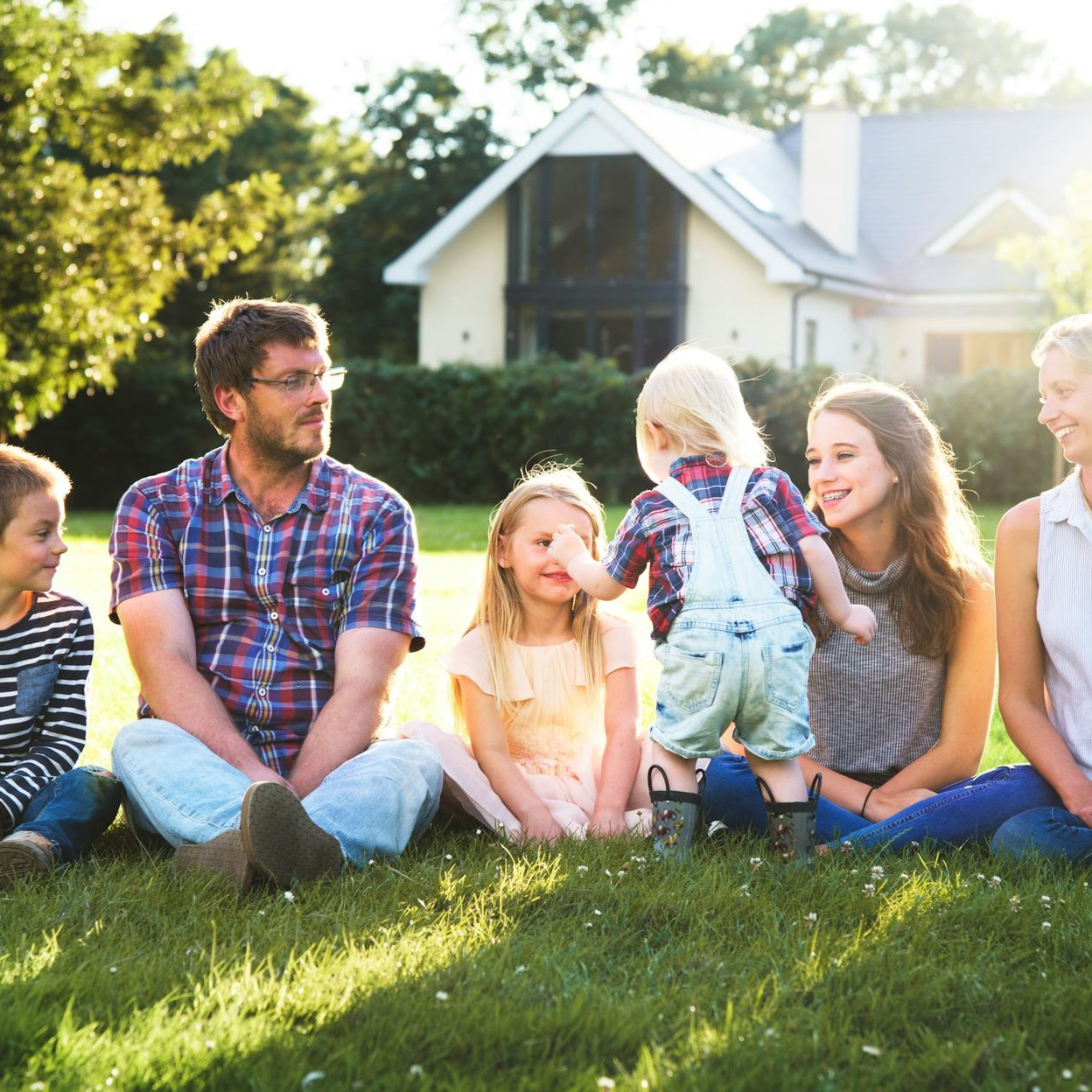 Family sitting on the floor smiling at a toddler. Mom holding child. Dad holding child's hand. Living room setting.
