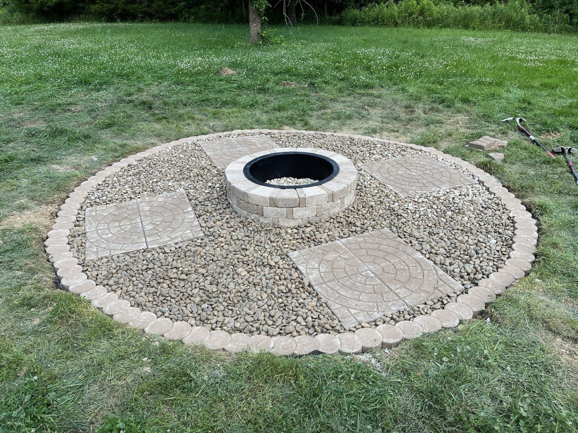 A fire pit is surrounded by gravel and bricks in the middle of a lush green field.