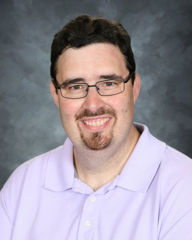 Man with glasses and goatee smiles, wearing a light purple polo shirt against a gray background.