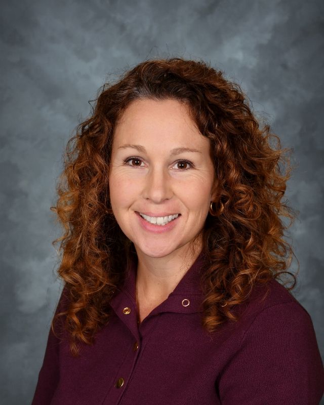Woman with auburn curly hair, smiling, wearing a dark burgundy shirt, against a mottled gray background.