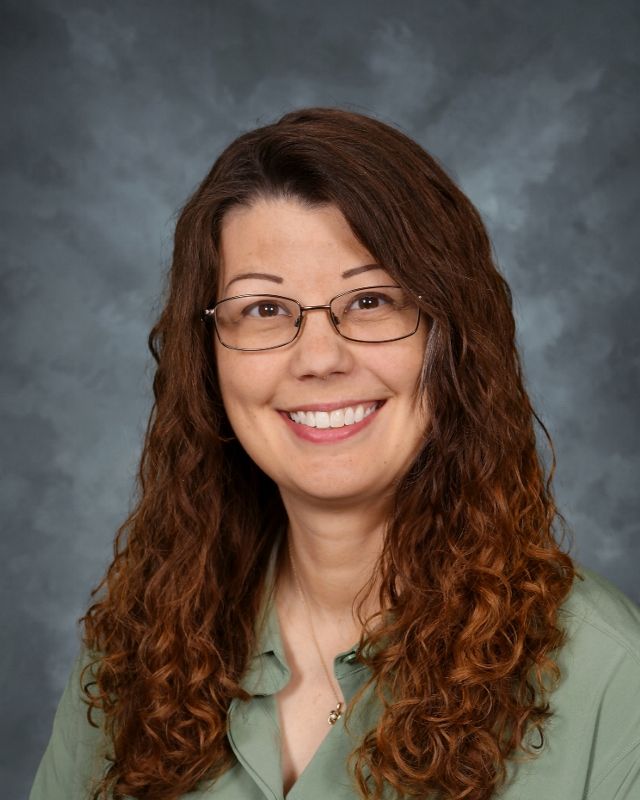 Woman with long curly brown hair and glasses smiling, wearing a green shirt.