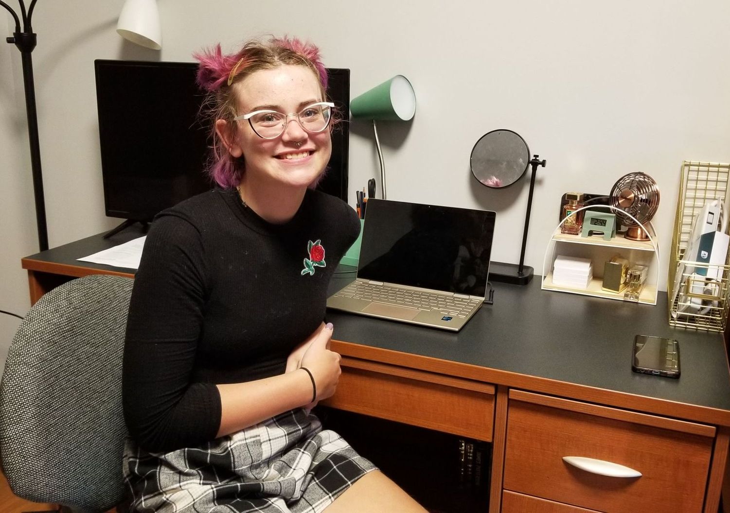 Woman smiling at a desk with a laptop, monitor, lamp, and decorations.