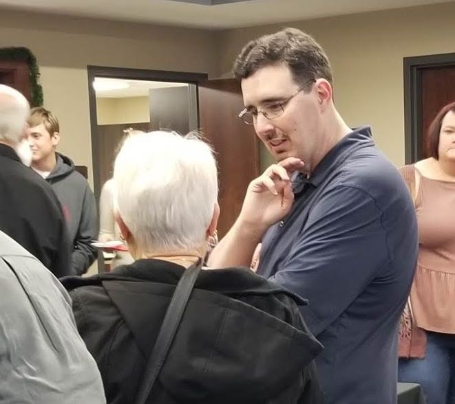 Man in glasses talks to woman with white hair; others in background.
