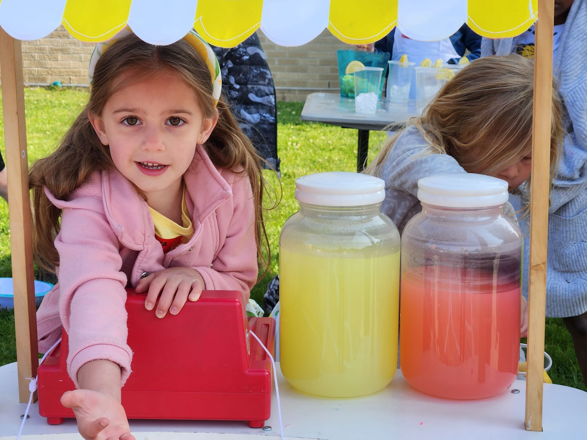 Girl at lemonade stand, smiling and gesturing. Jars of yellow and pink lemonade.