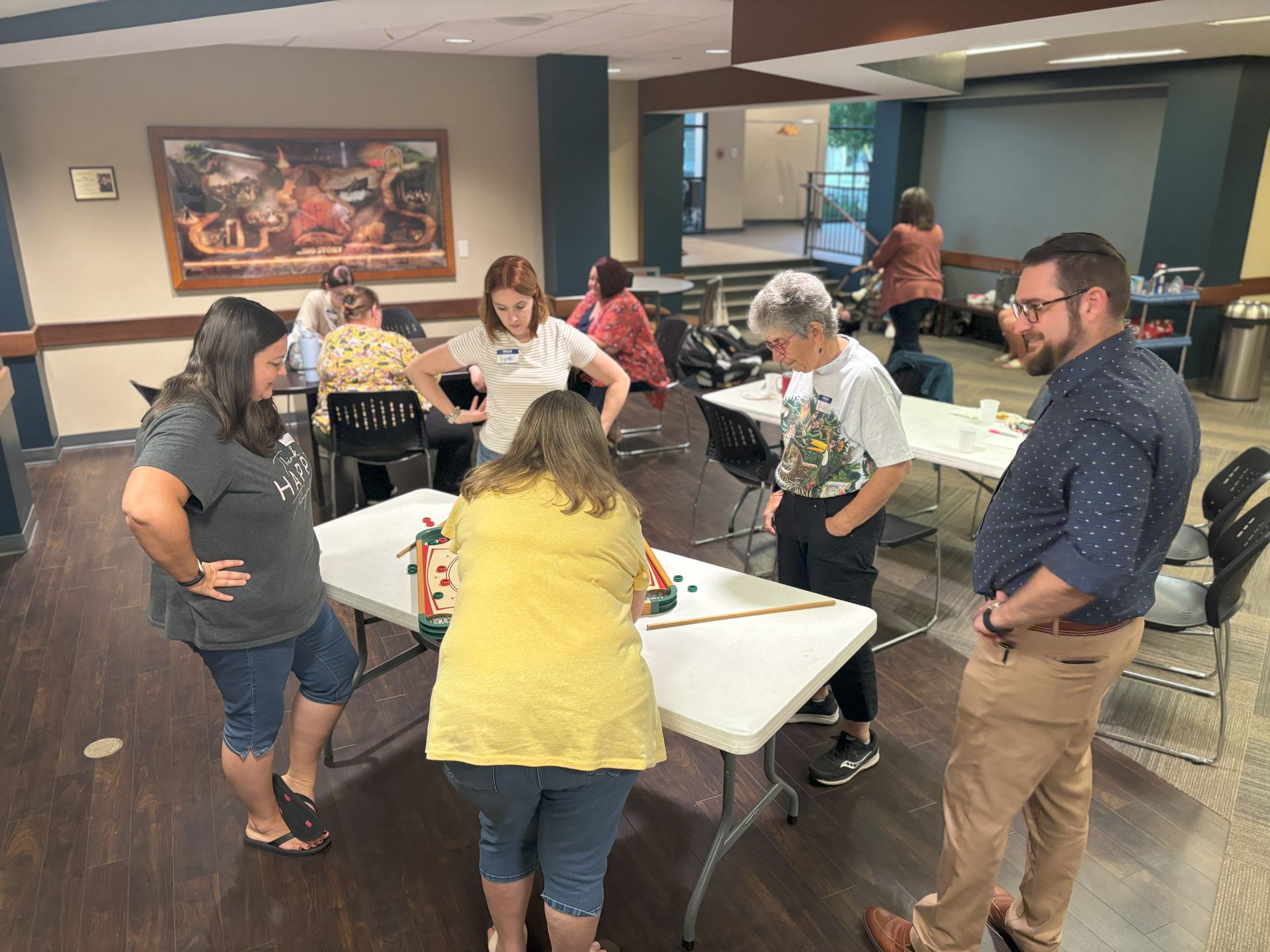 People gathered around tables, engaging in an activity indoors. Various expressions and gestures displayed.