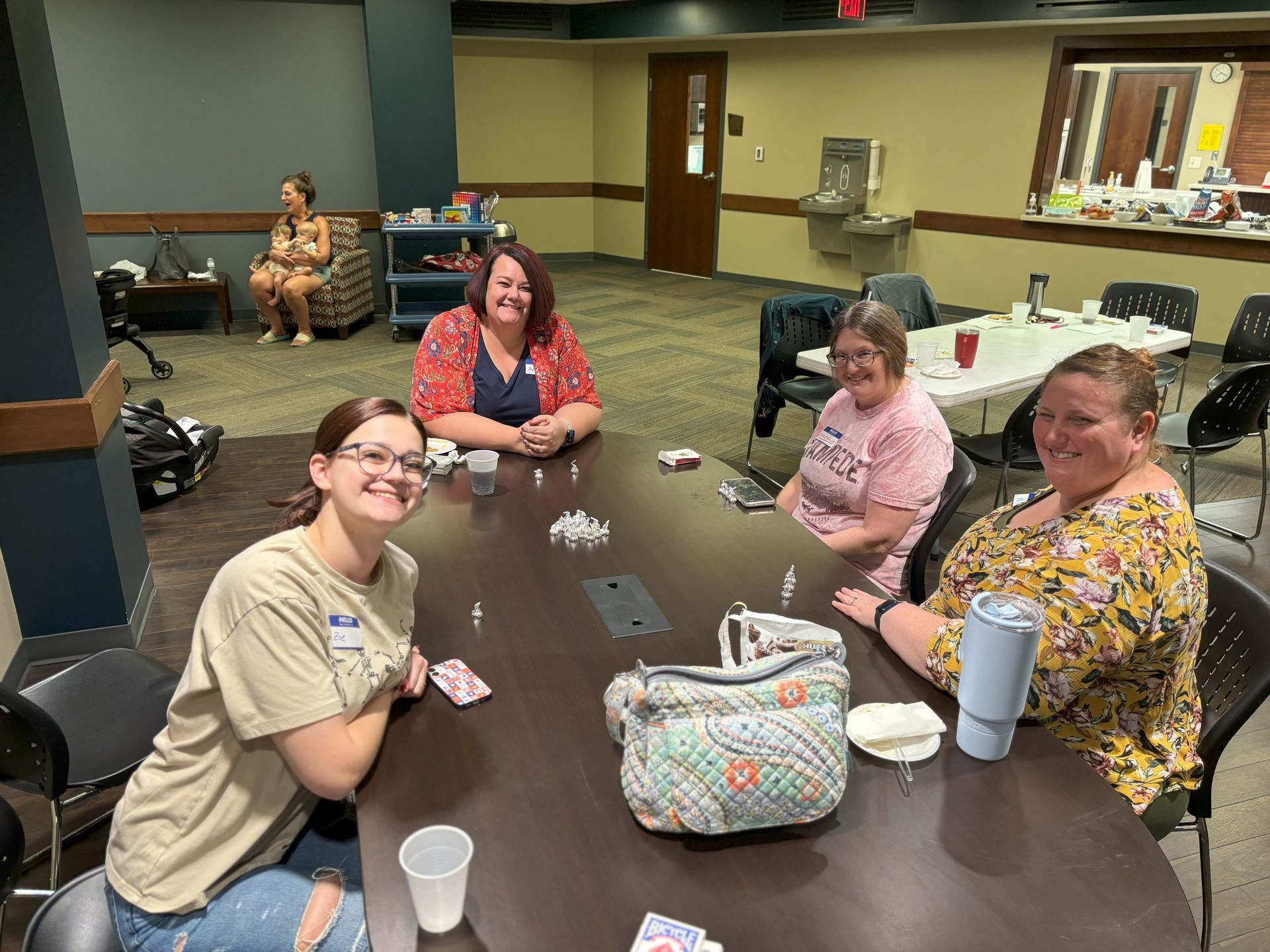 Four women at a table smiling, playing cards. One woman sits in a chair in the background. Indoor setting.