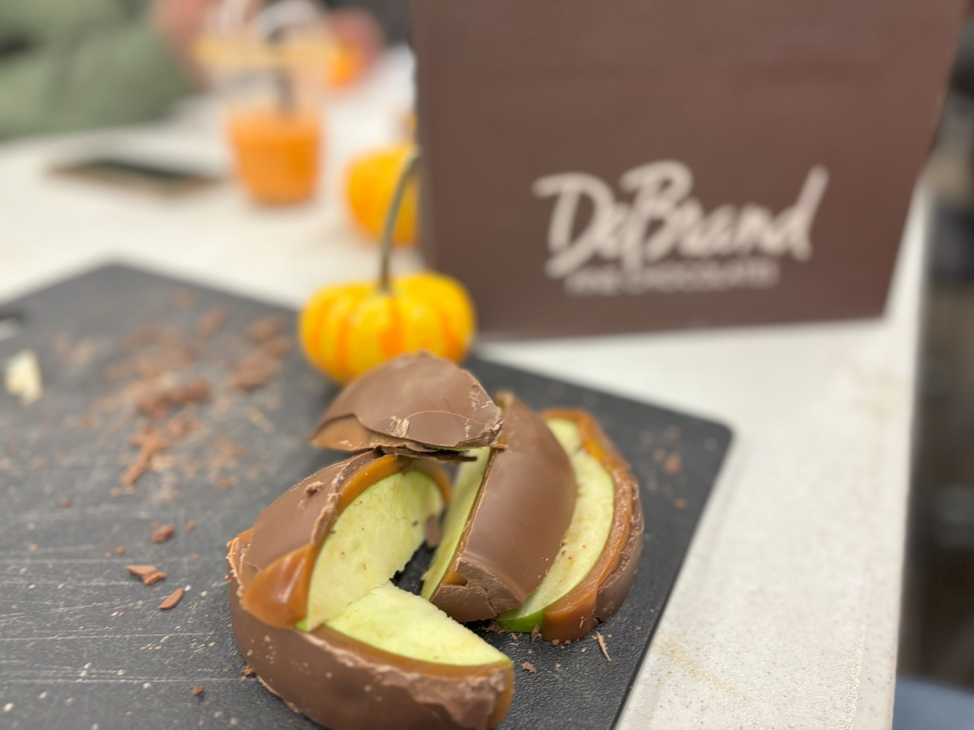 Chocolate-covered apple slices on a cutting board, with a small pumpkin and a DeBrand box in the background.