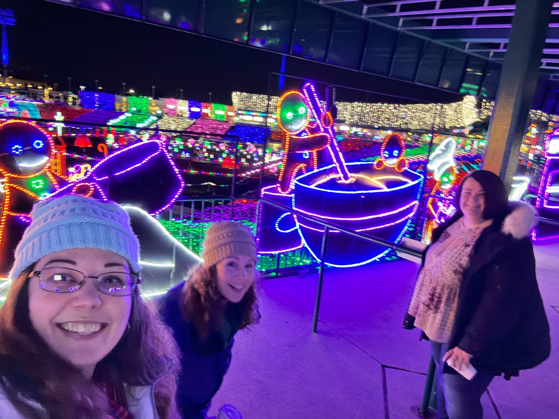 Three smiling women pose in front of bright holiday light display featuring gingerbread people and a coffee cup.
