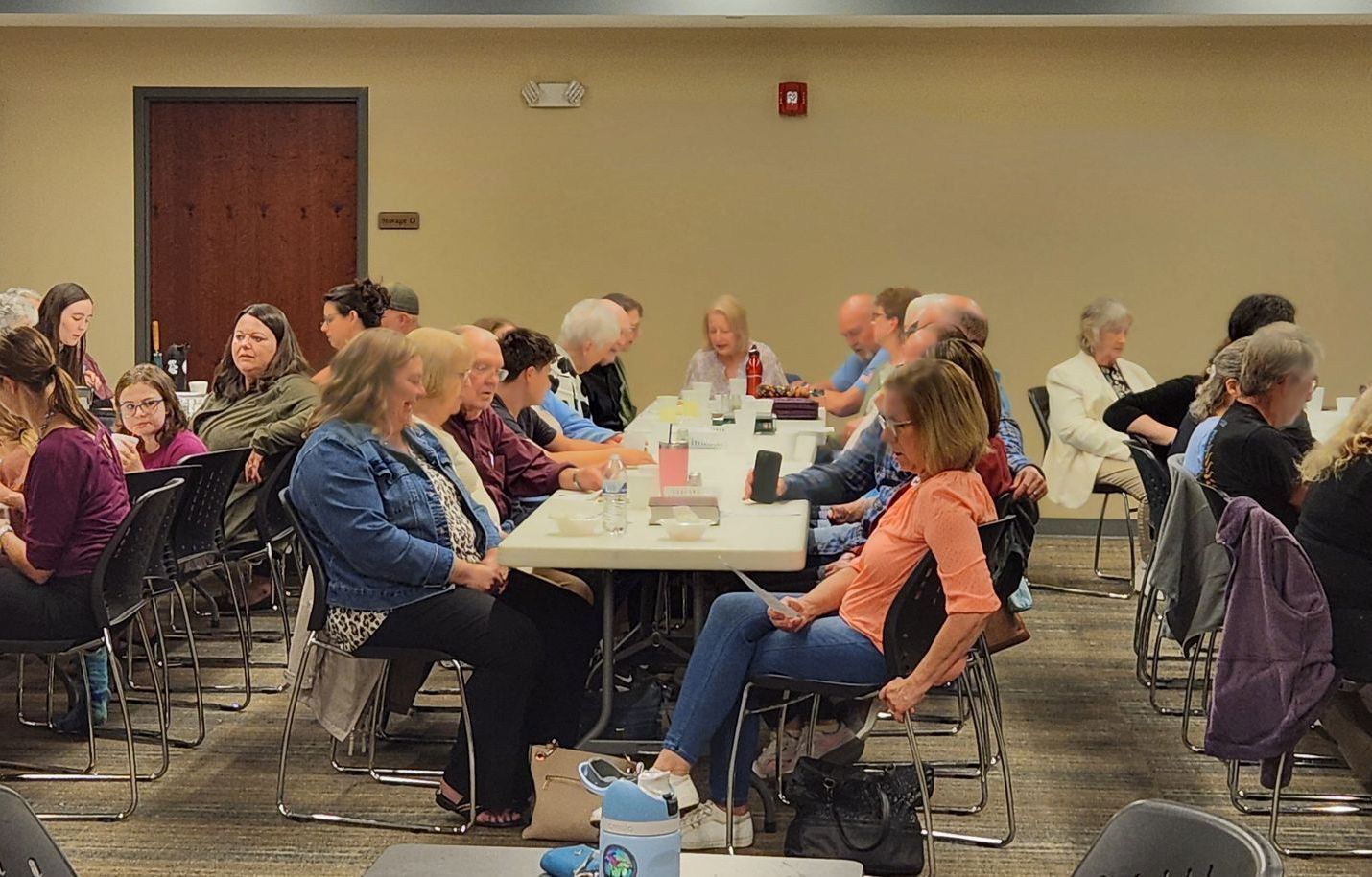 People seated at tables in a brightly lit room; some are talking, others are listening.