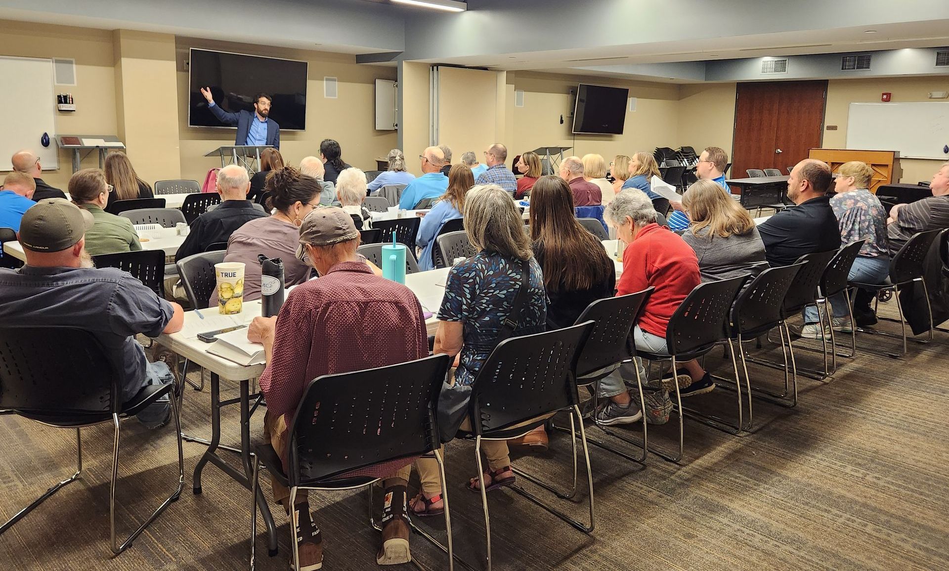 A man gestures to an audience seated at tables in a conference room.