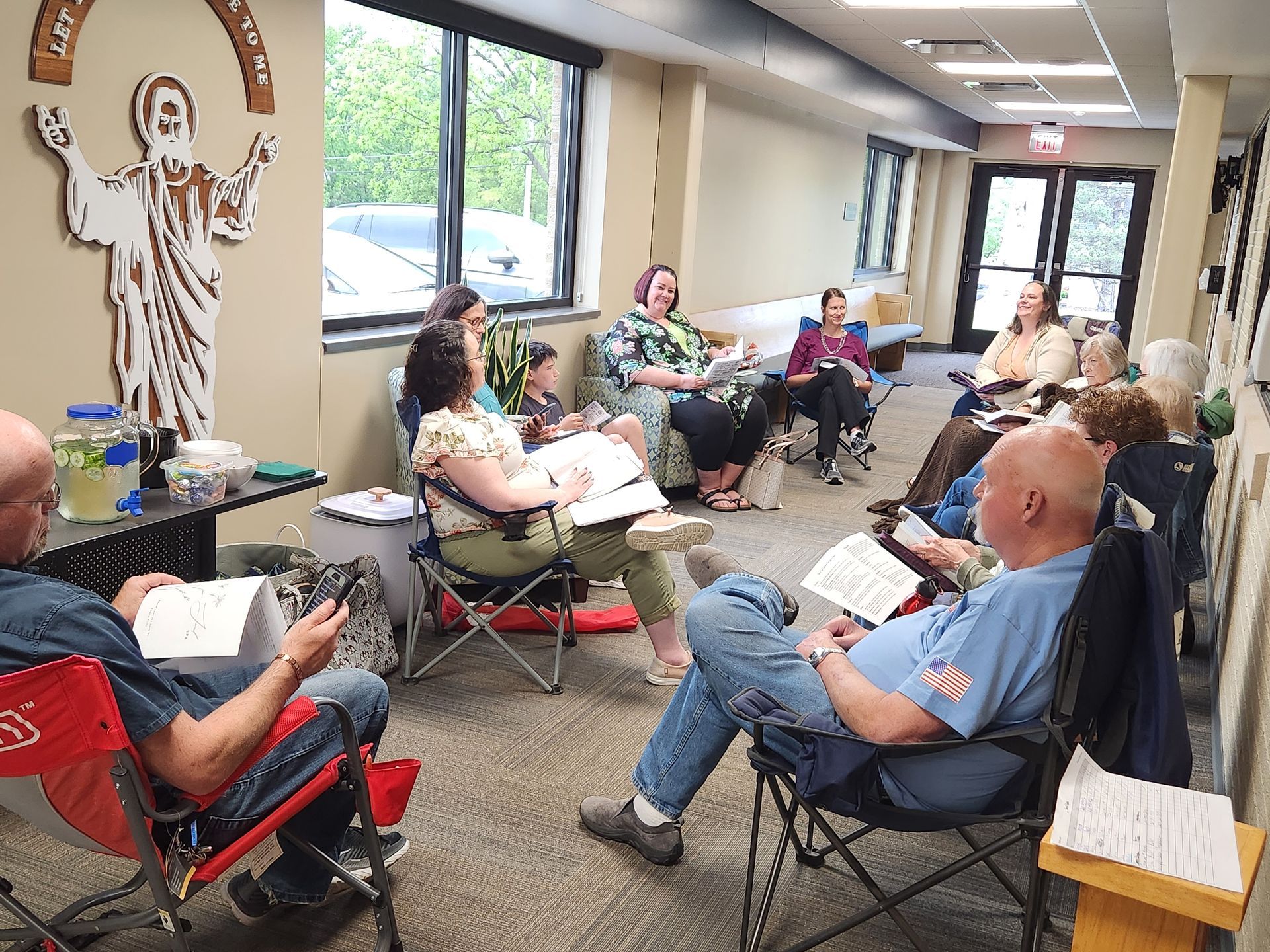 Group of people sitting in chairs in a room, engaged in conversation. A religious symbol is on the wall.