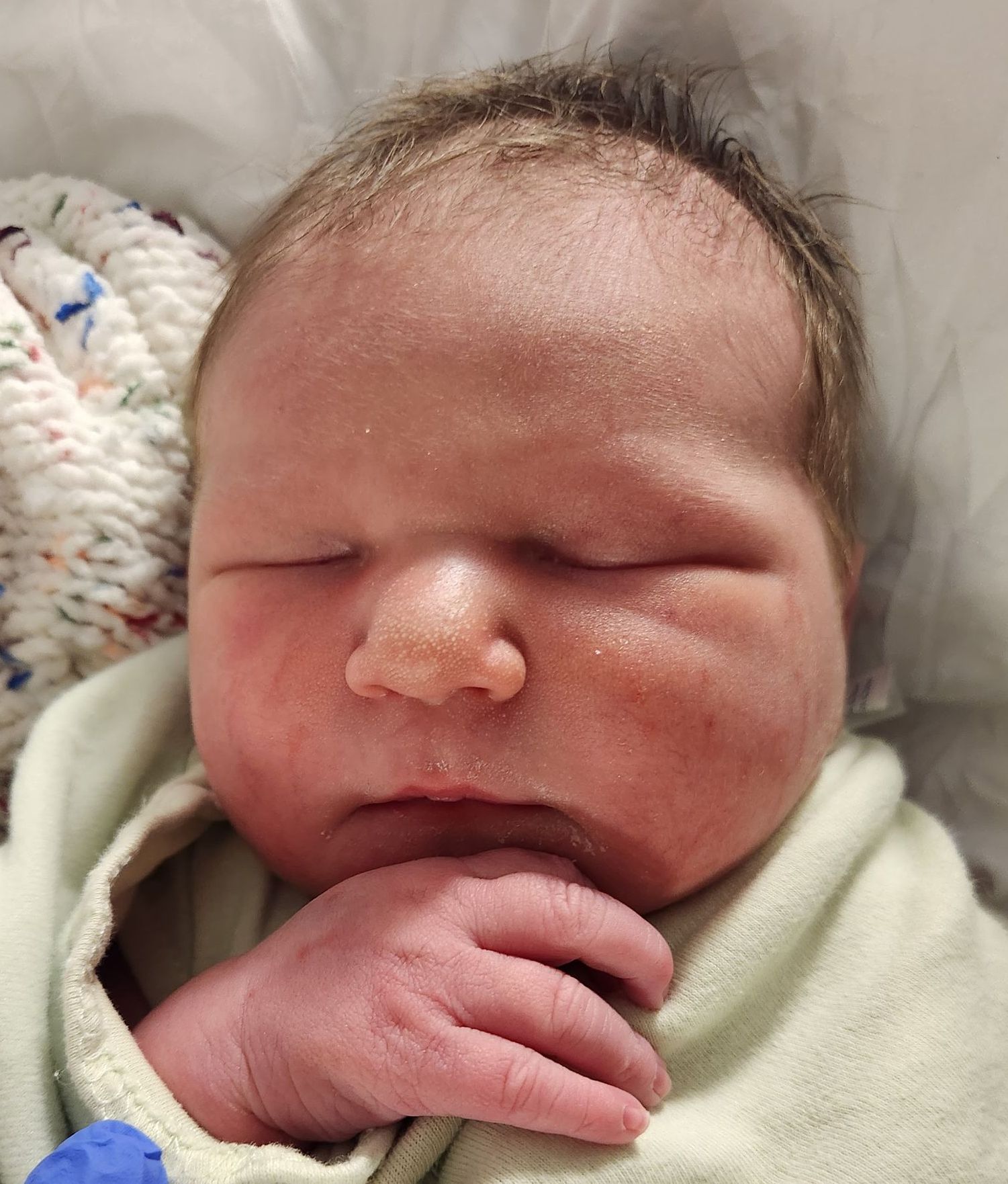 Newborn baby swaddled in green, sleeping peacefully; red cheeks and small hand visible.