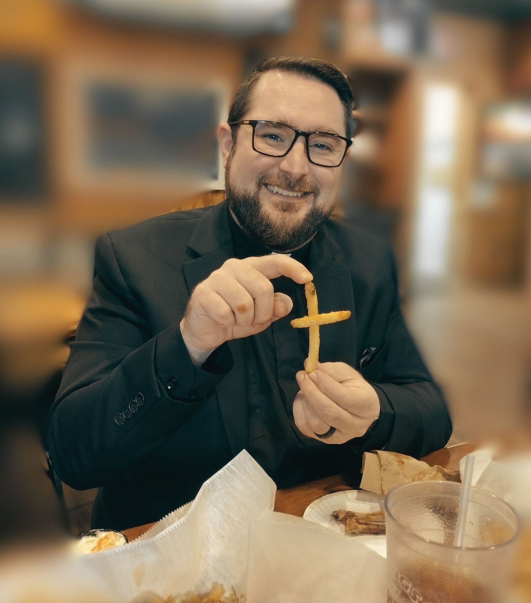 Man in black clerical attire smiles, holding French fries arranged in a cross shape at a restaurant.