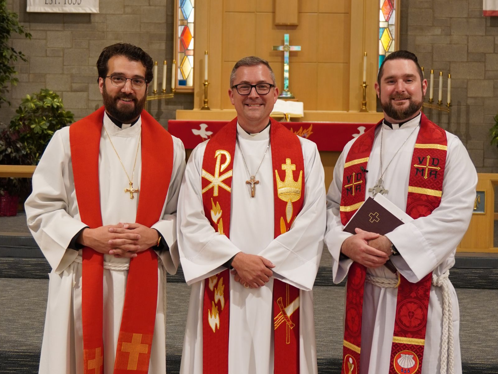 Three clergy members stand in a church. They wear white robes and red stoles, smiling.