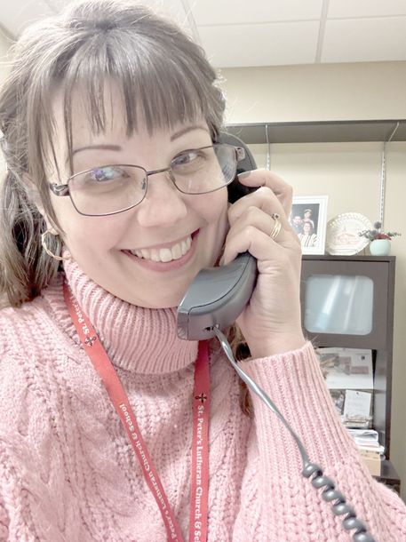 Woman with glasses smiles while holding a telephone receiver. She wears a pink sweater in an office setting.