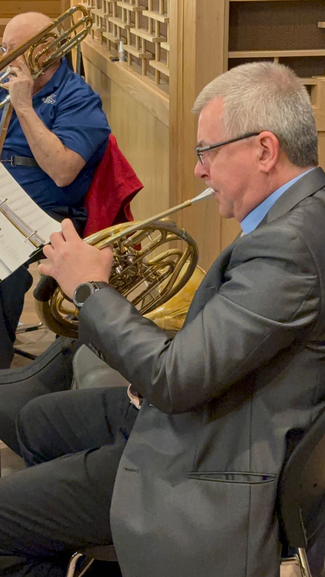 Man playing a French horn in a seated position, another person playing a trombone in the background.