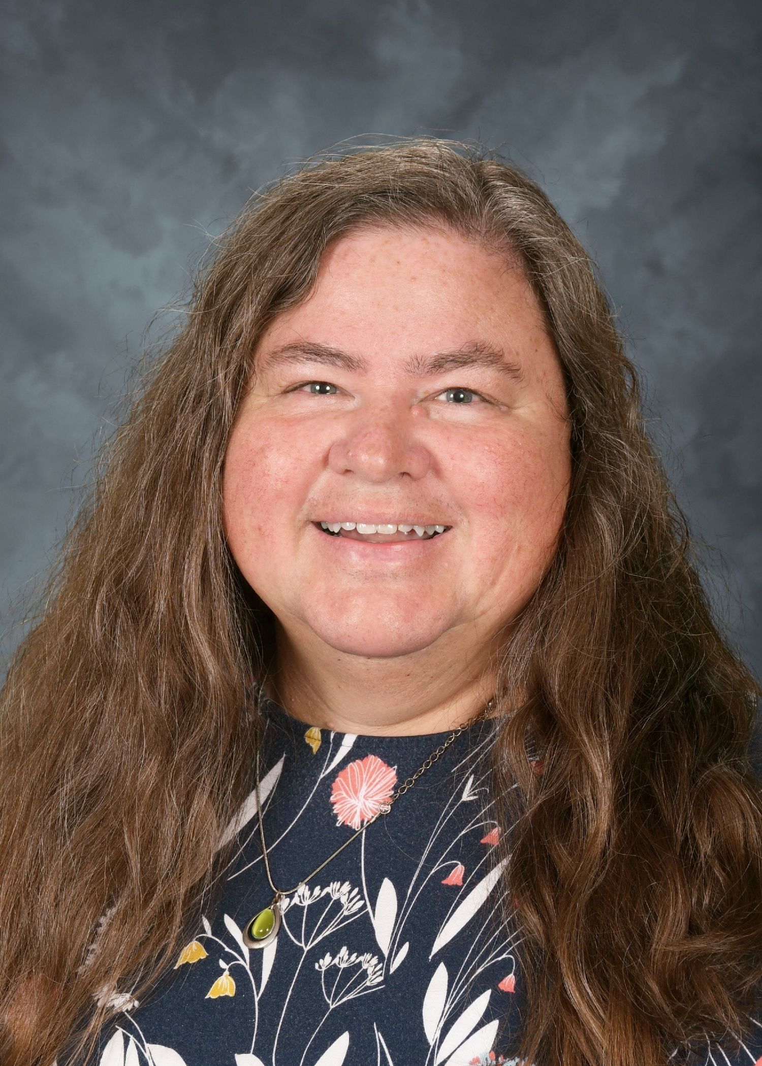 Woman with long, wavy brown hair, smiling, wearing a floral patterned top against a mottled blue background.
