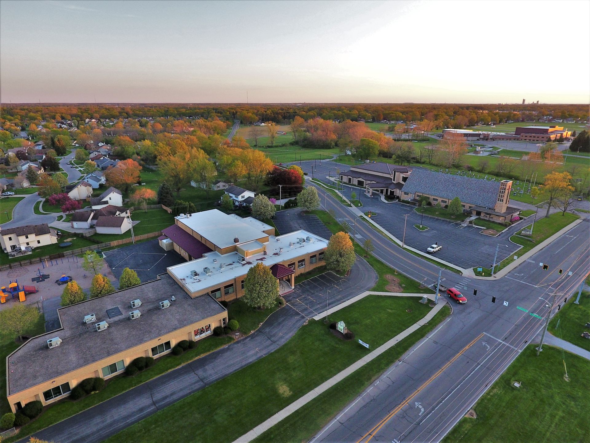 Aerial view of buildings, roads, and a residential area at dusk. Trees display fall foliage with a pink and orange sky.