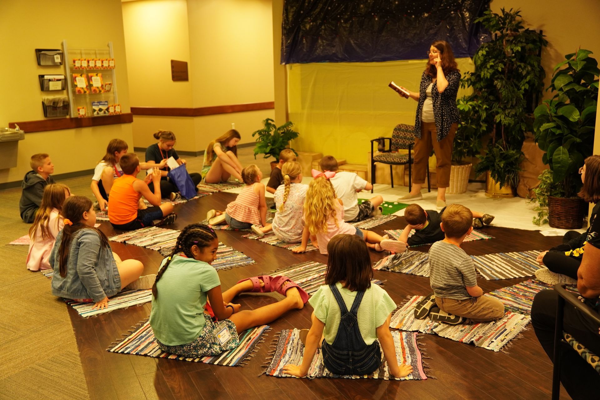 Children seated on mats listen to a woman reading in a room decorated with plants.
