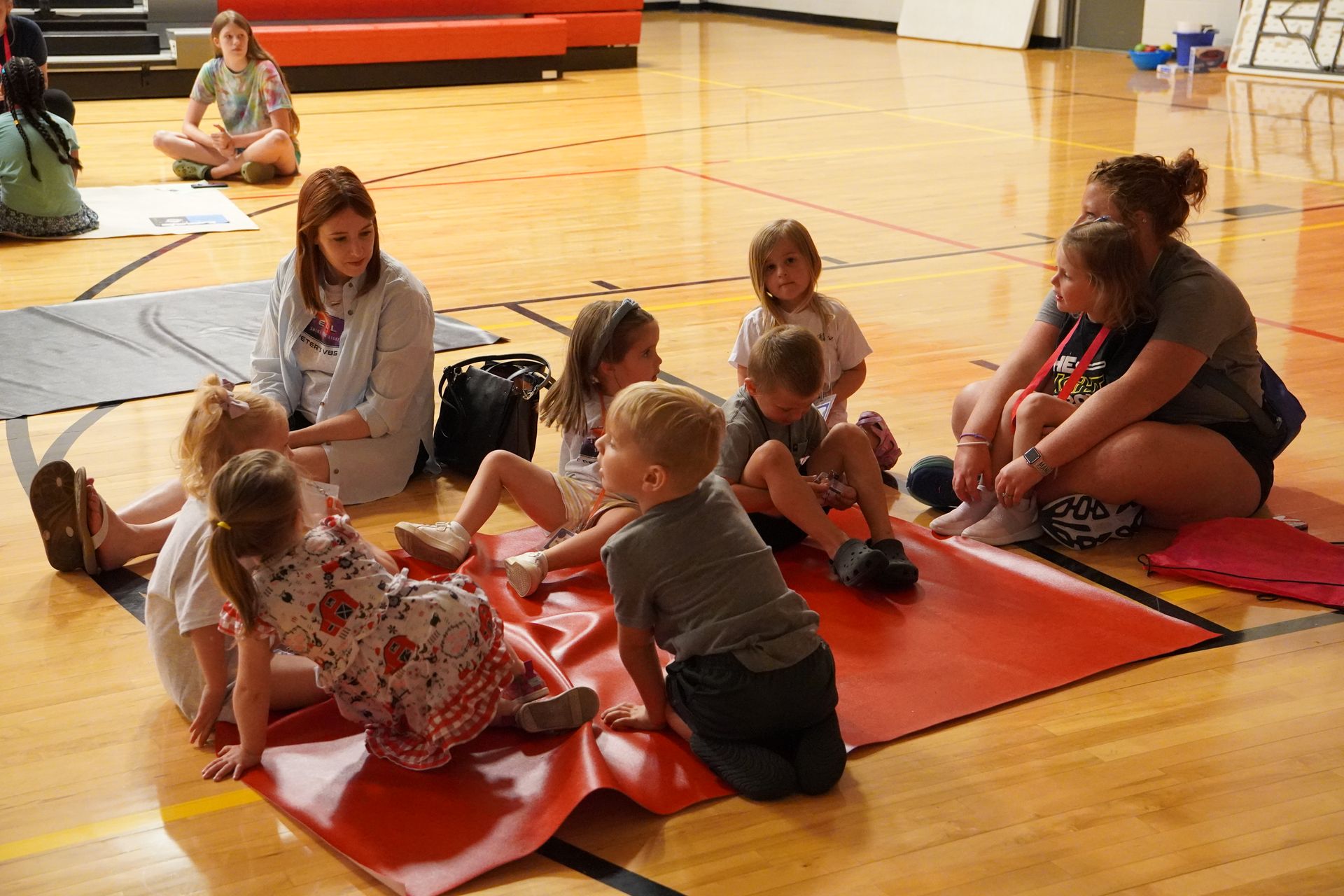 Children and adults sitting on mats in a gymnasium. One adult appears to be reading to the children.