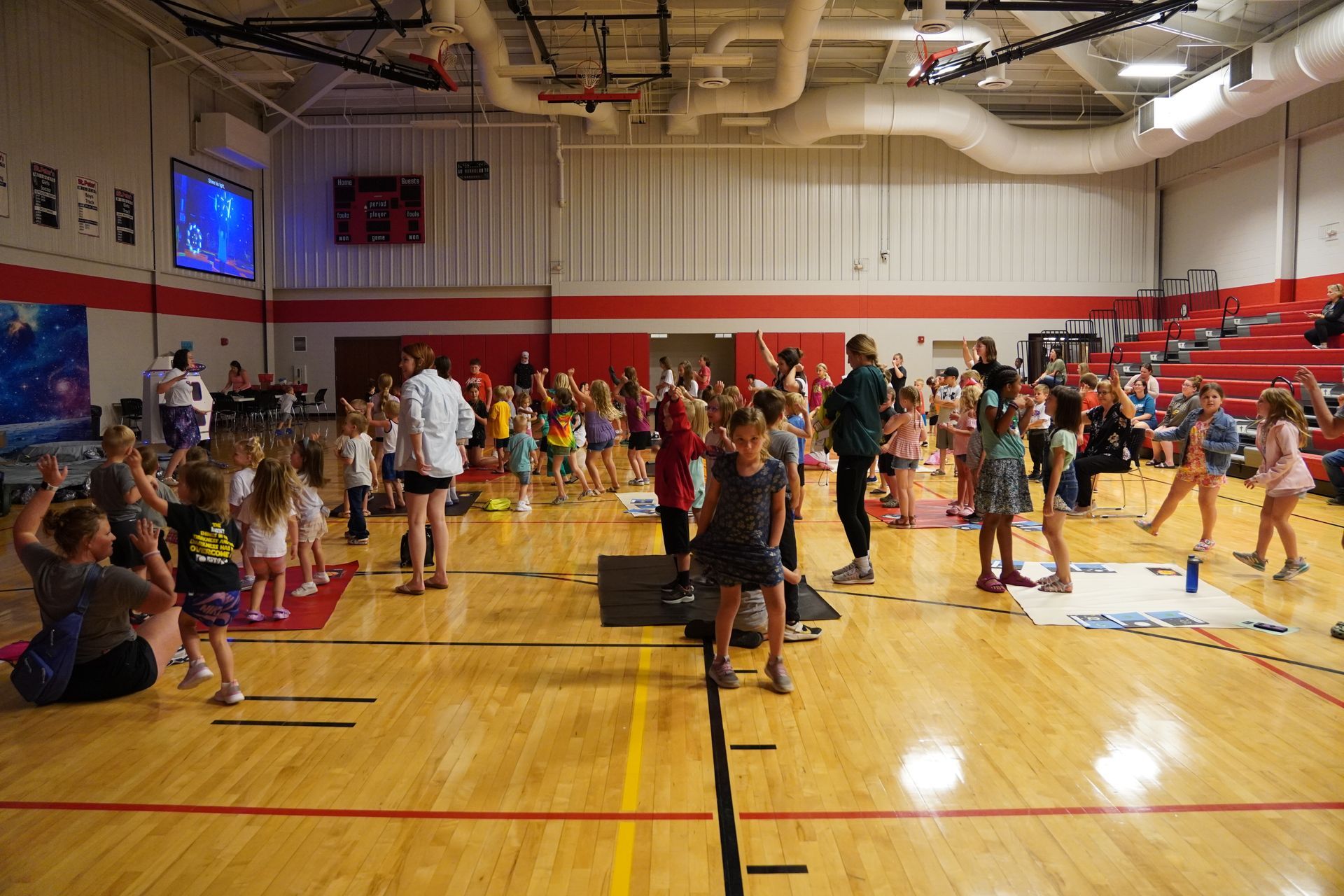 Gymnasium with children dancing, adults supervising. Wooden floor, red trim, and bleachers in background.