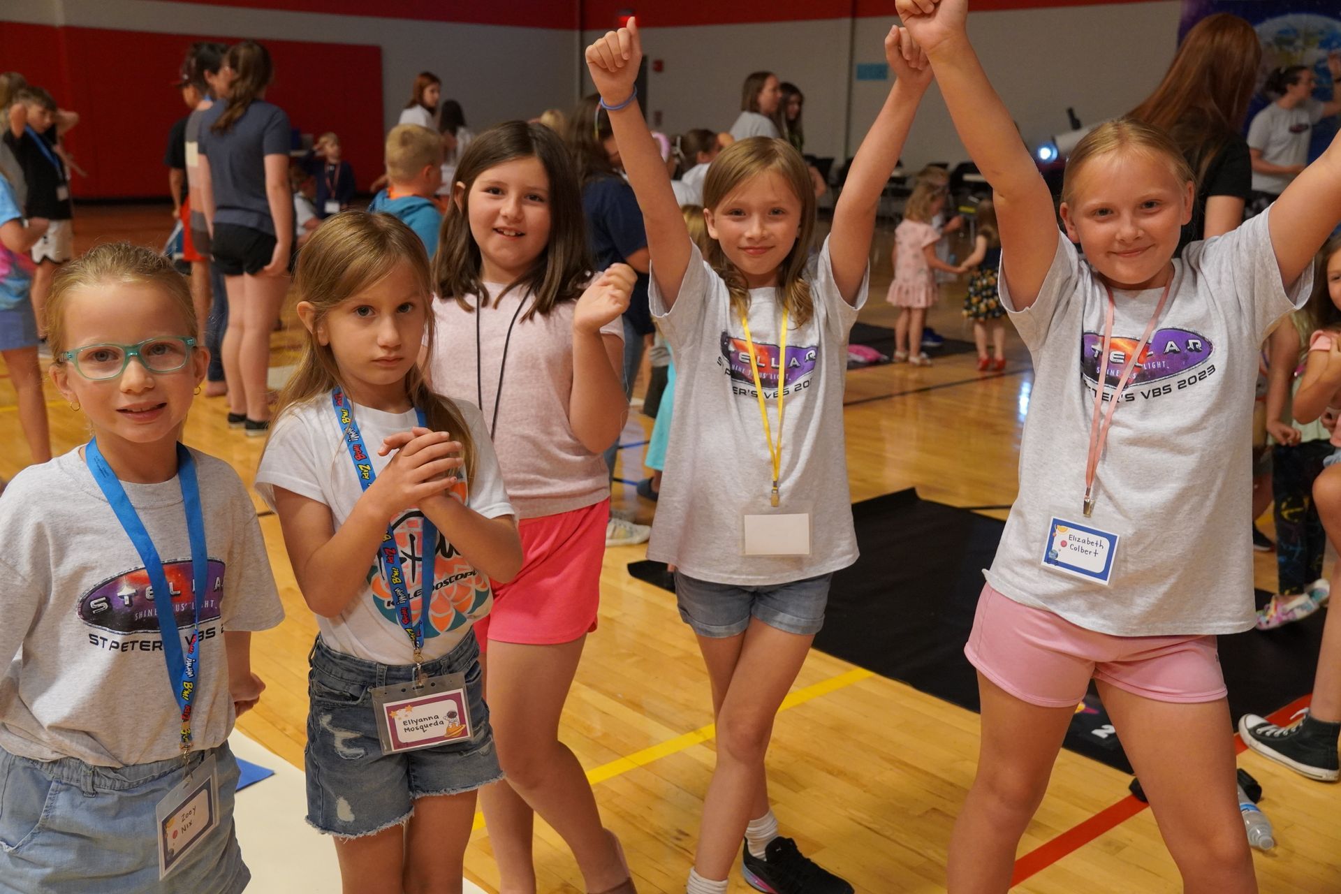 Five young children in a gymnasium, some with arms raised. They wear light-colored shirts and shorts.