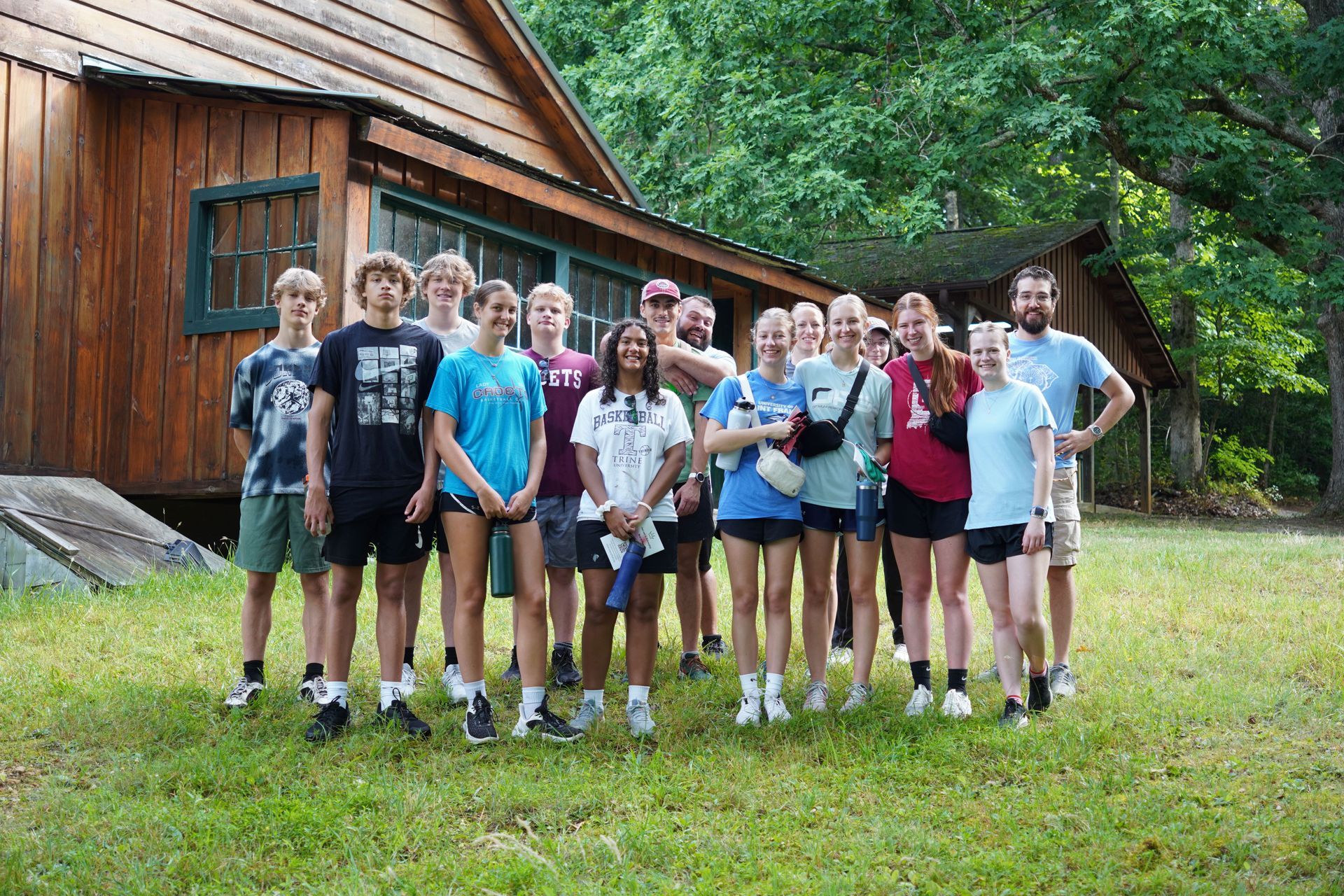 Group of people standing in front of a wooden cabin in a grassy area, some wearing t-shirts.