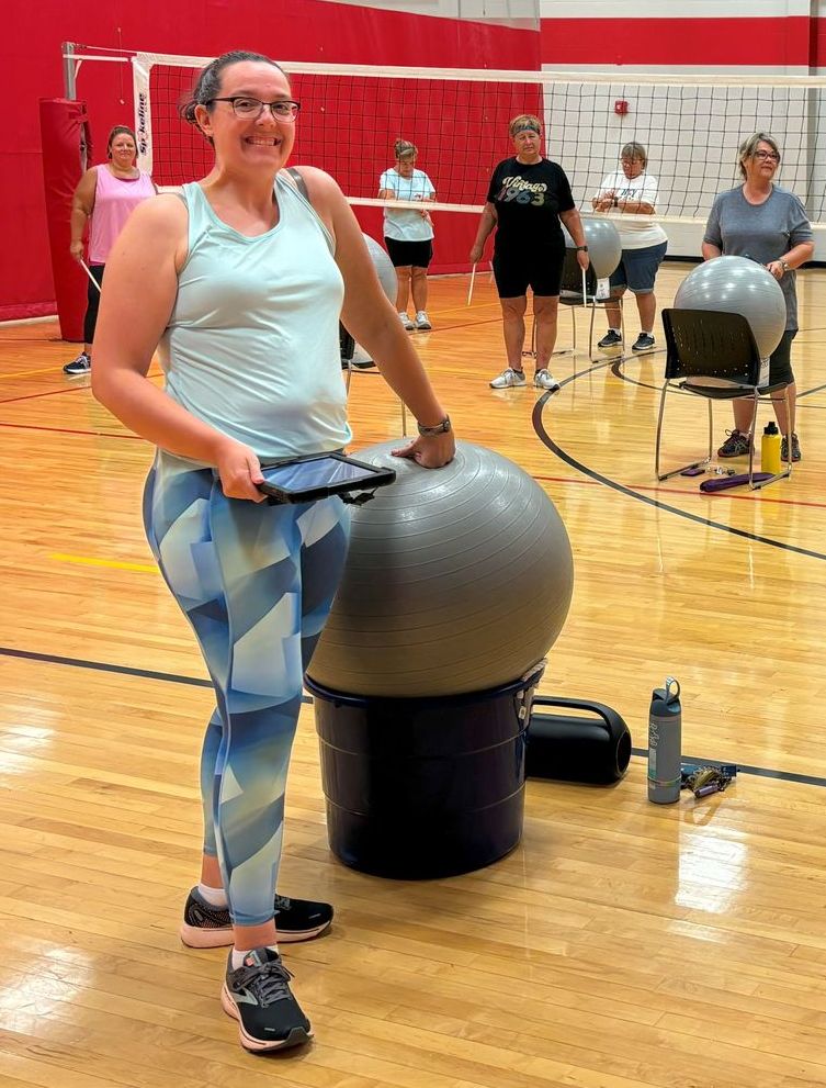 Woman in gym clothes with tablet, next to exercise ball; group of people in the background.