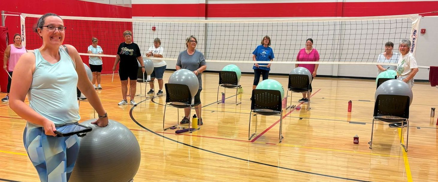 Woman leading a group fitness class in a gymnasium. Participants are exercising with chairs and exercise balls.