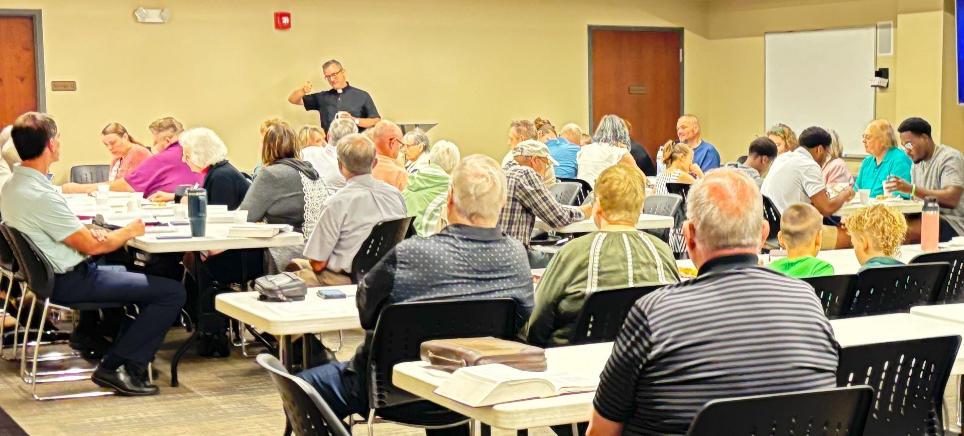 Group of people in a meeting room, listening to a Pastor Danny Eggold speaking. Tables and chairs are visible. Bible Study