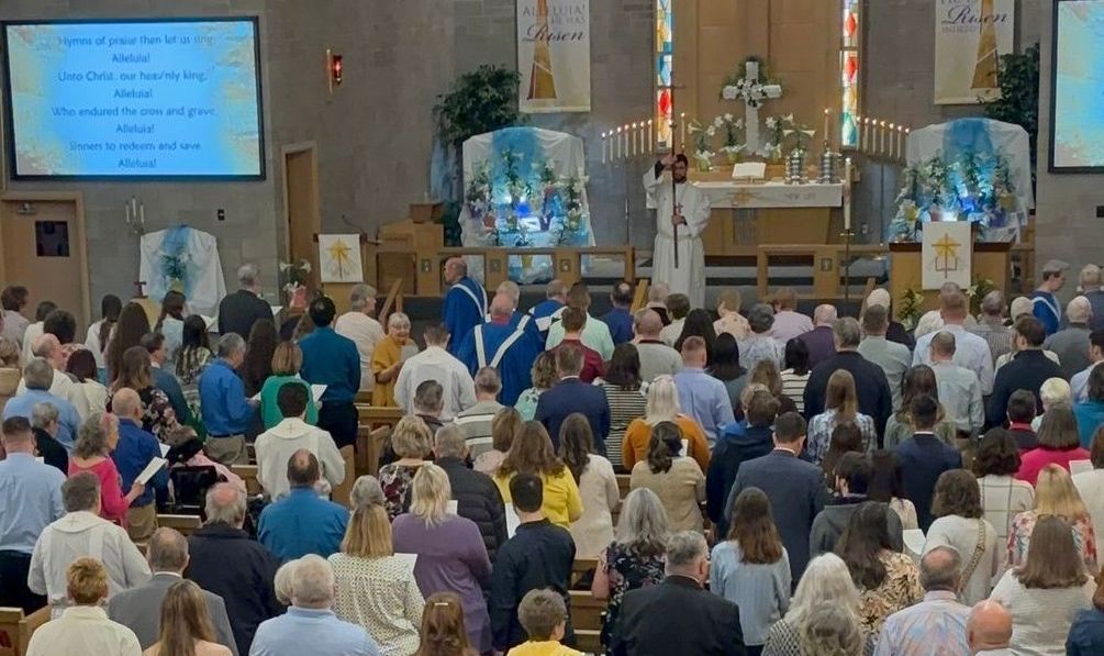 Worship service in a church, with a crowd of people. A service is in progress near an altar. Blue and white decorations are present.