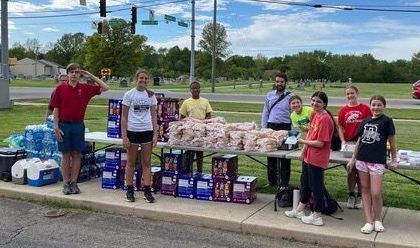 People stand by table of food and drinks outdoors, assisting in a food drive.
