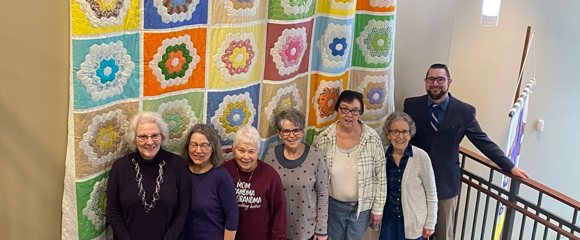 Group of people posing by a colorful quilt. A man in a suit stands behind them on stairs.
