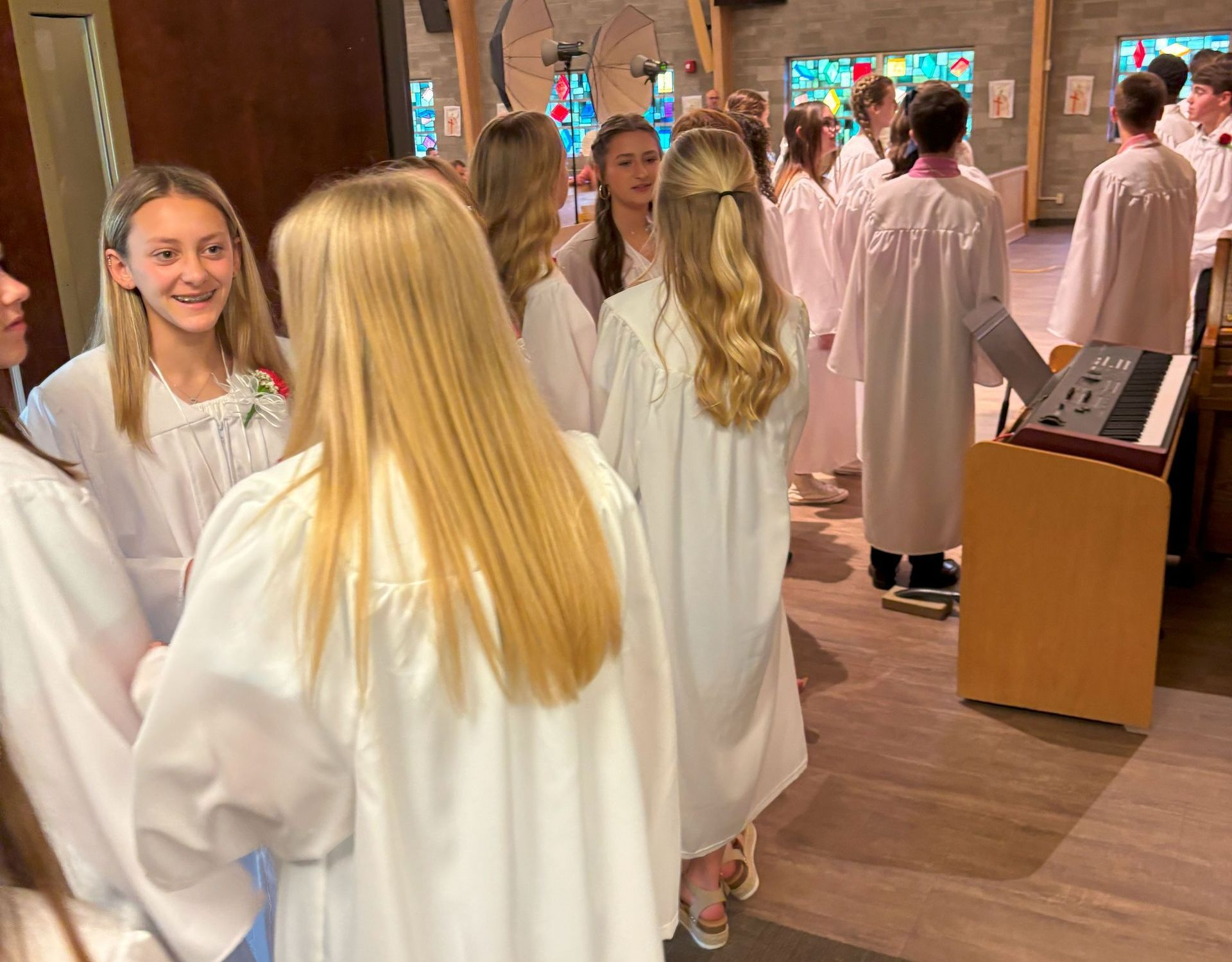 Children in white robes stand together inside a church. Some are smiling, preparing for a ceremony.