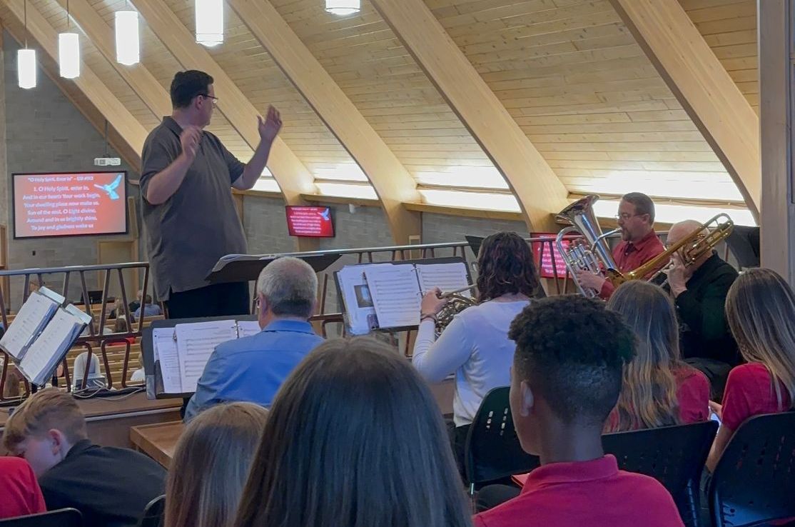 Band conductor leading musicians playing instruments in a wooden-beamed building.
