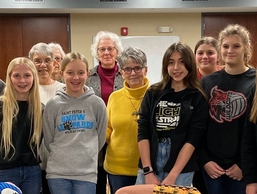 Group of women and girls smiling, posing for a photo indoors. Some wear sweatshirts; neutral background.