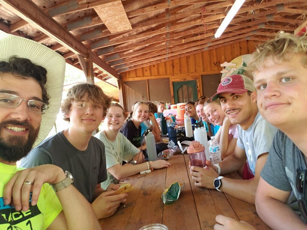Group of people smiling around a wooden table under a covered outdoor structure.