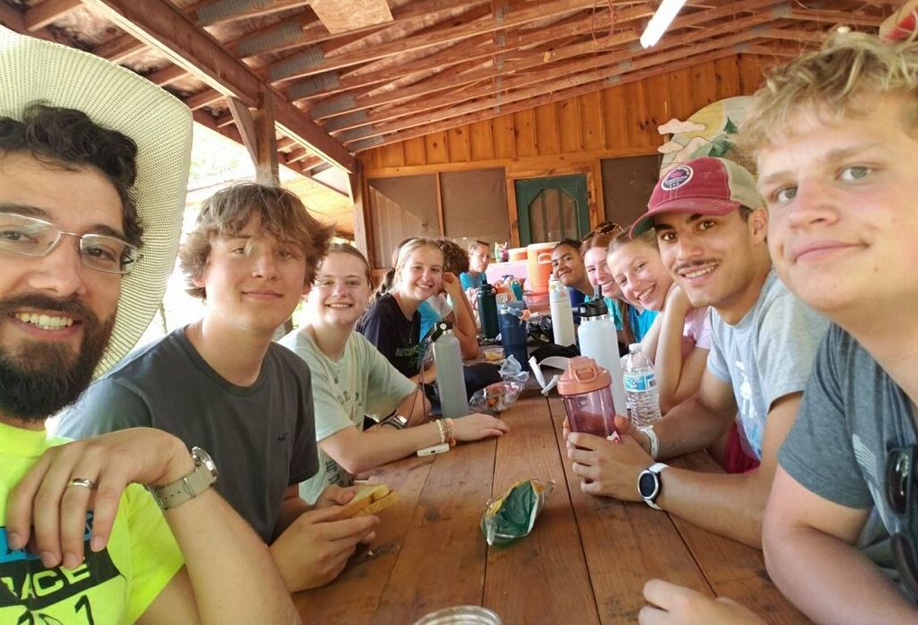 Group of people smiling around a long wooden table outdoors under a covered structure.