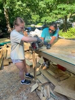 Two people cutting wood with a circular saw outdoors, surrounded by wood scraps.