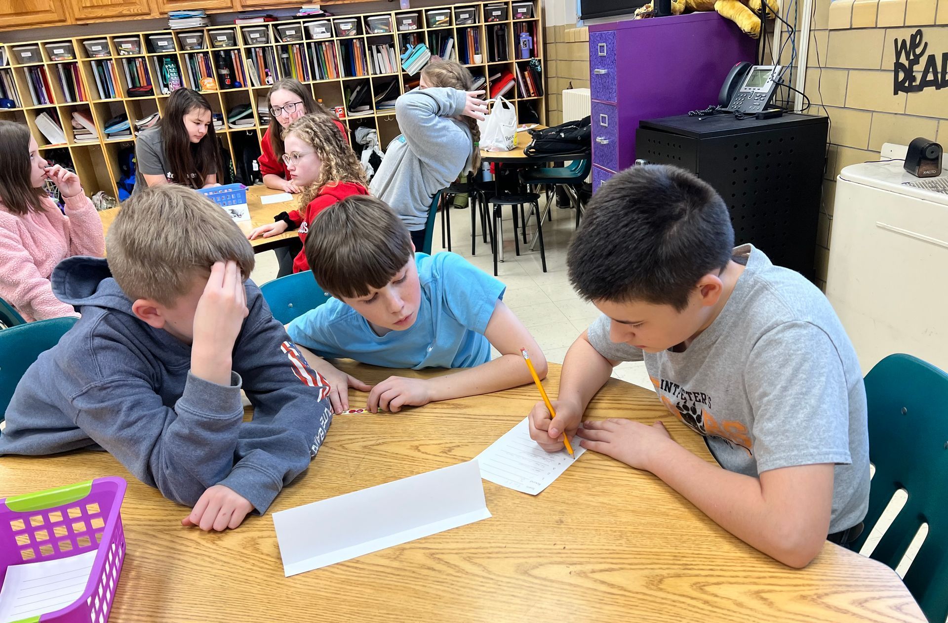 Students seated around a table, writing on paper in a classroom.