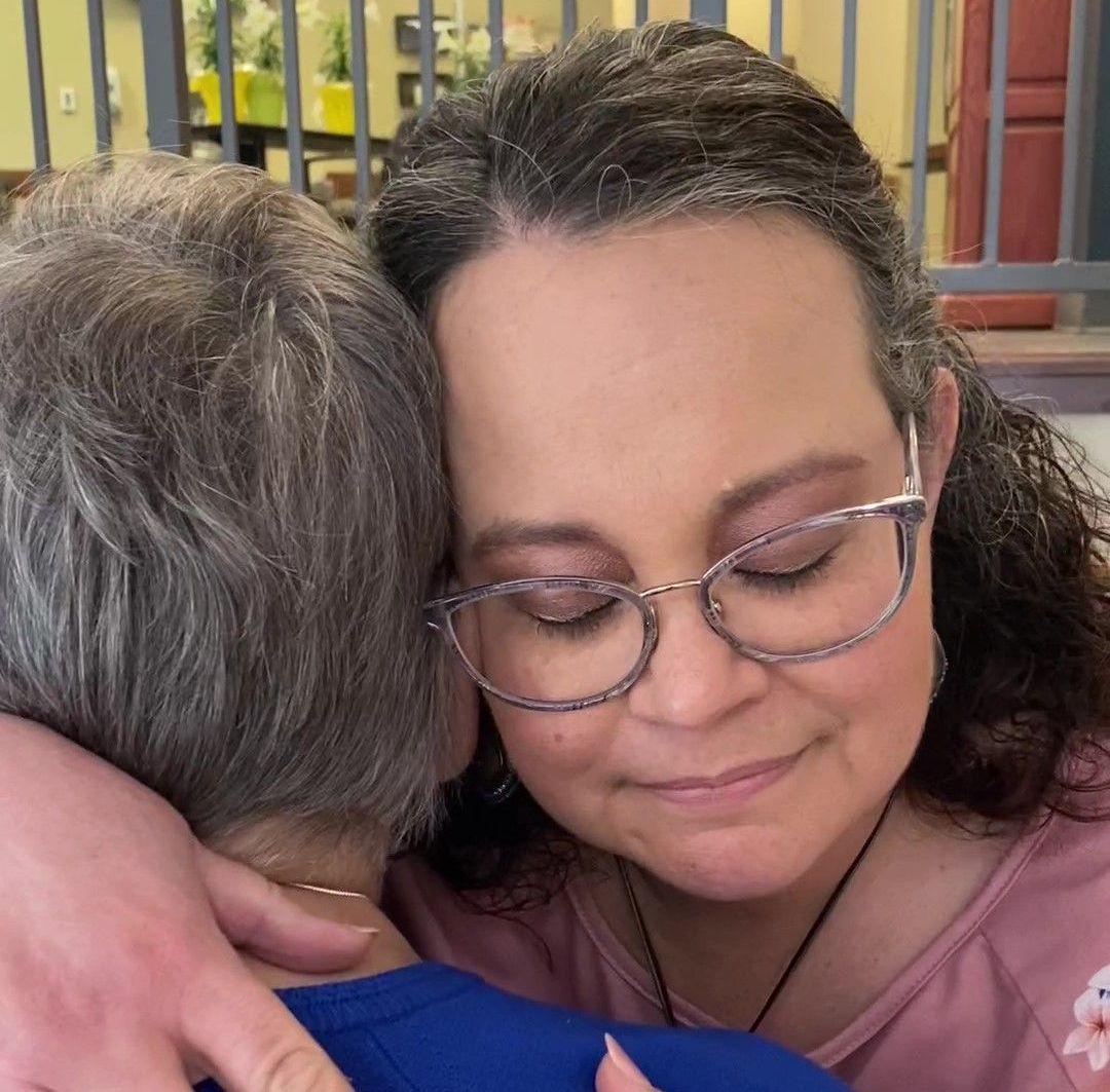 Woman hugs another person, both with eyes closed. Indoors, warm lighting.