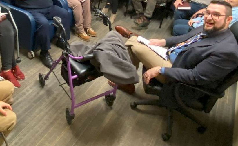 Man in a suit smiles, seated with legs crossed, resting on a walker in a room with others.