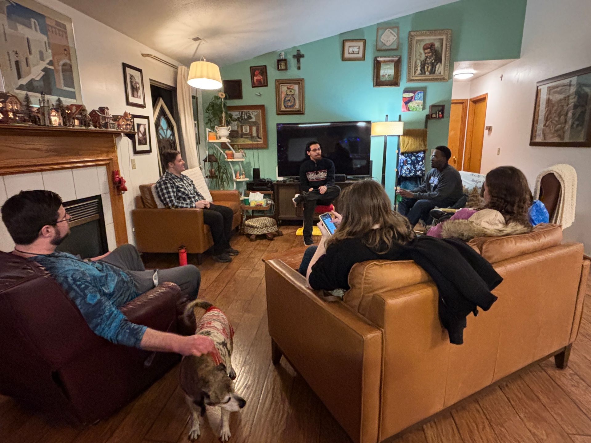 Group of people in living room, discussing. Several sitting on leather couches, with dog. Fireplace, art on walls.