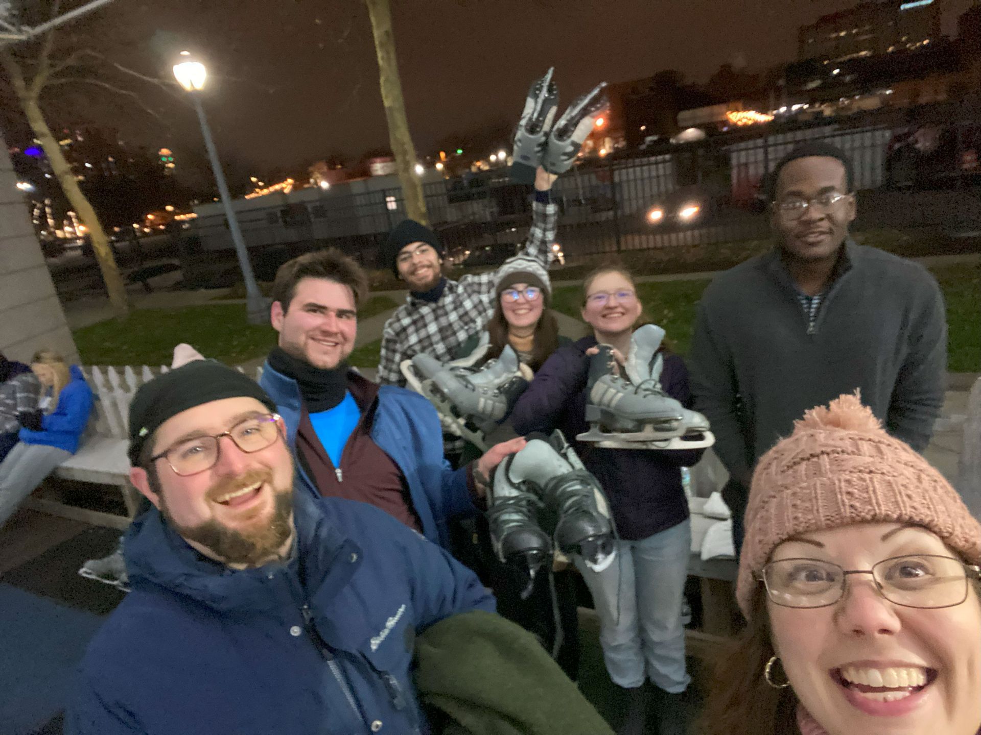Group of friends smiling at an ice skating rink at night; several hold ice skates.