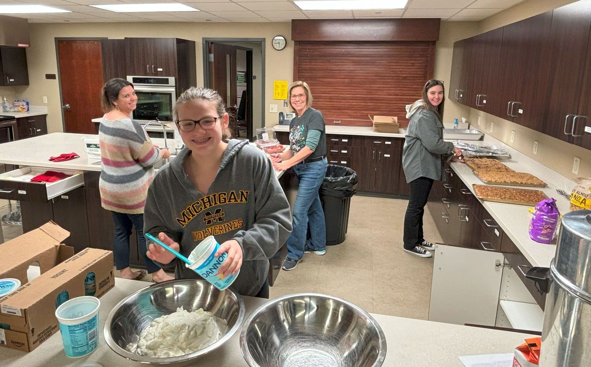People preparing food in a kitchen. Woman pours from a container into a bowl. Others arrange items on a counter.
