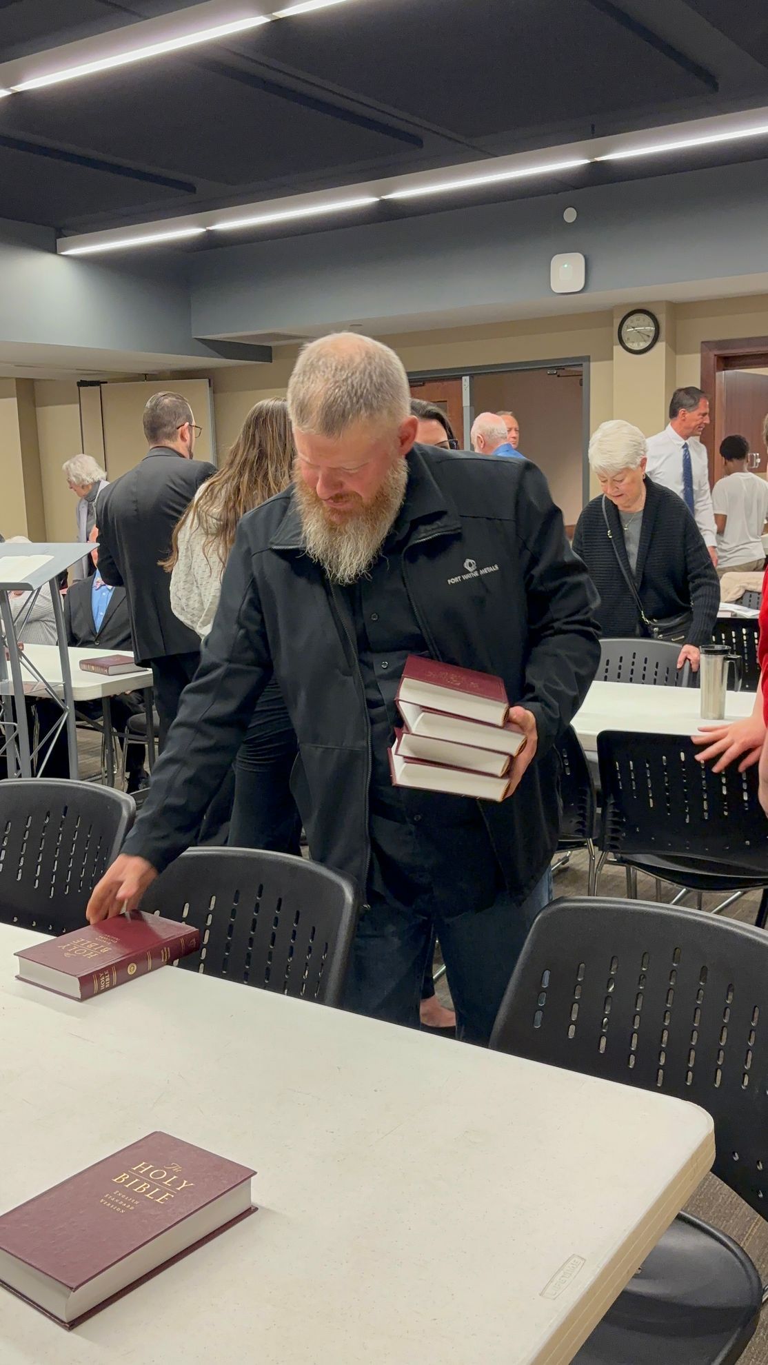 Man with a beard placing books on a table in a room with other people.