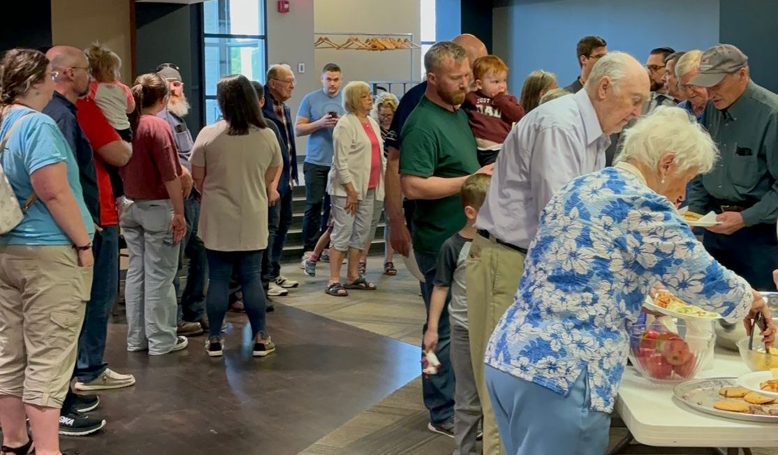 People at a gathering, some at a table with food, others stand in a line. Indoors, neutral colors.