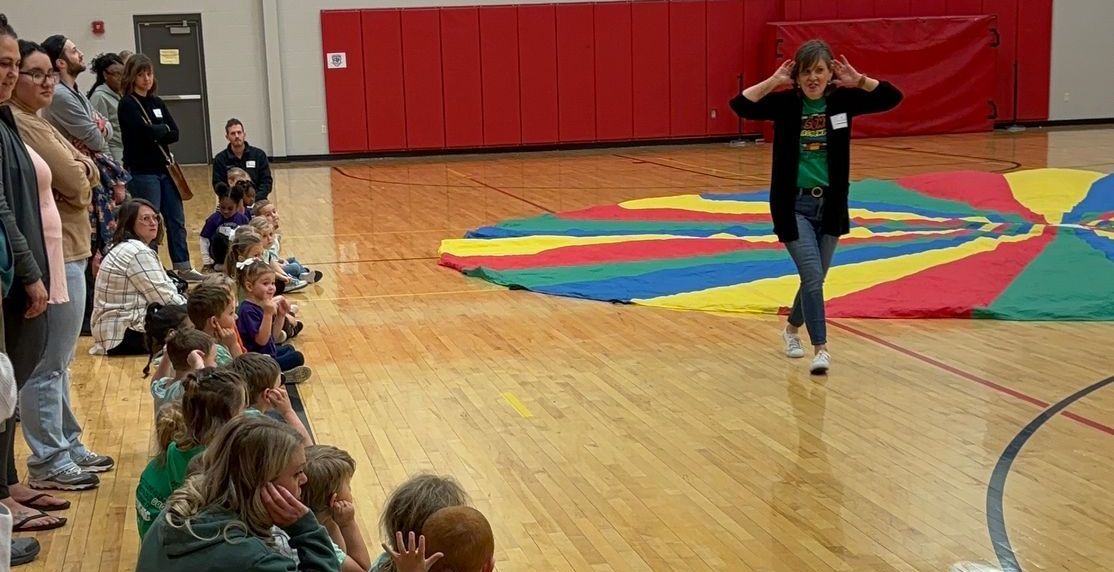 A woman with hands on her head walks on a gym floor; children and adults watch near a colorful parachute.
